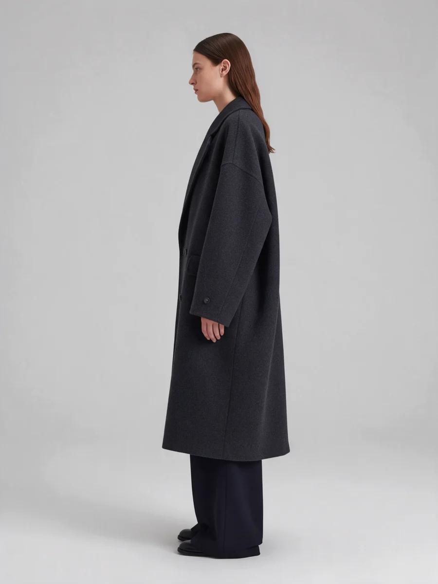 Model in an oversized charcoal wool coat and wide trousers on a light grey studio background