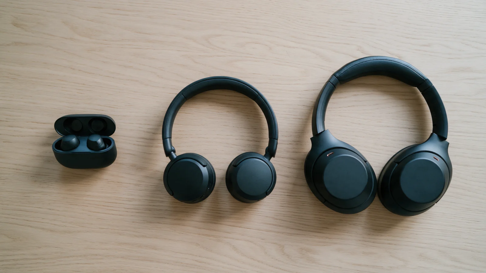 Wireless earbuds in a case, on-ear headphones and over-ear headphones side by side on an oak table